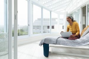Senior woman using laptop on bed in bright sunroom, enjoying leisure time, copy space. Elderly, technology, relaxing, senior lifestyle, computing, home environment